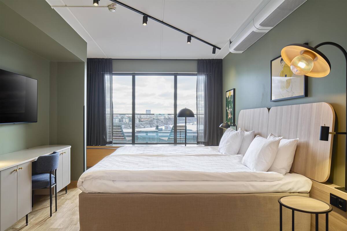 Une chambre d’hôtel moderne, dotée d’un grand lit, d’un bureau et offrant une vue sur la ville grâce à de larges fenêtres.