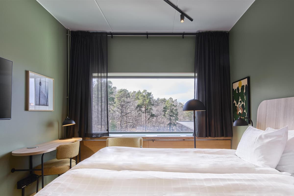 Une chambre d’hôtel avec un grand lit, un bureau, une chaise, une lampe et une fenêtre donnant sur des arbres.