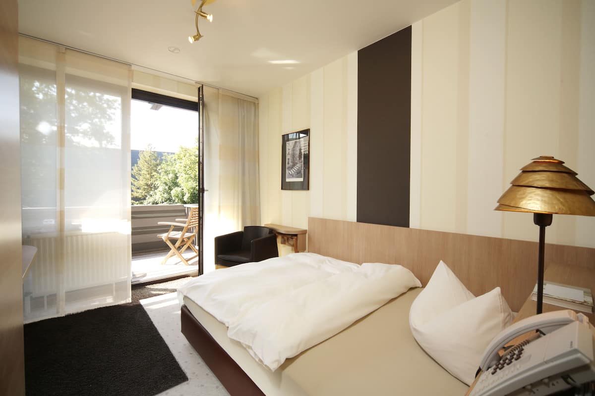 A hotel room with a bed, a desk, a chair, a lamp, and a view of the outdoors.