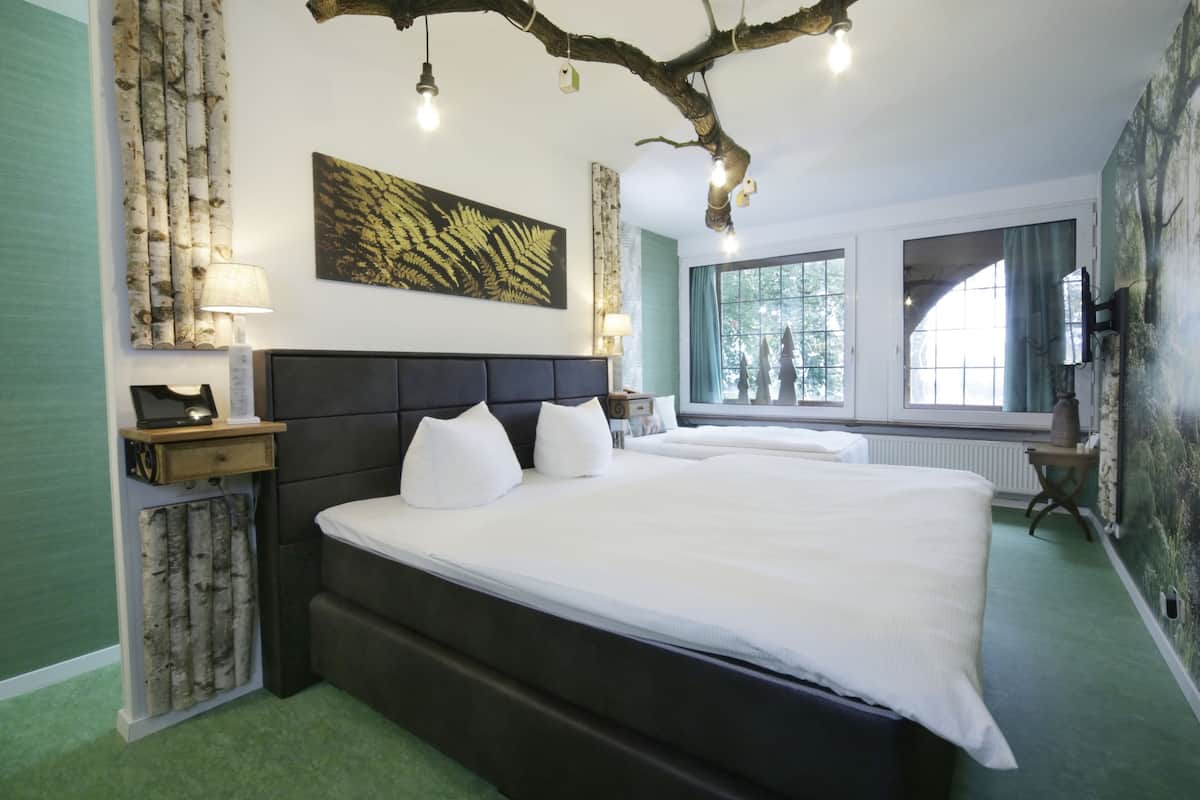 A bedroom with a large bed, a headboard, a nightstand, a lamp, a window with curtains, and a wall with a leaf pattern.