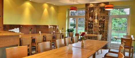 Een restaurant met houten tafels en stoelen, een accentmuur van steen en uitzicht op groen buiten.