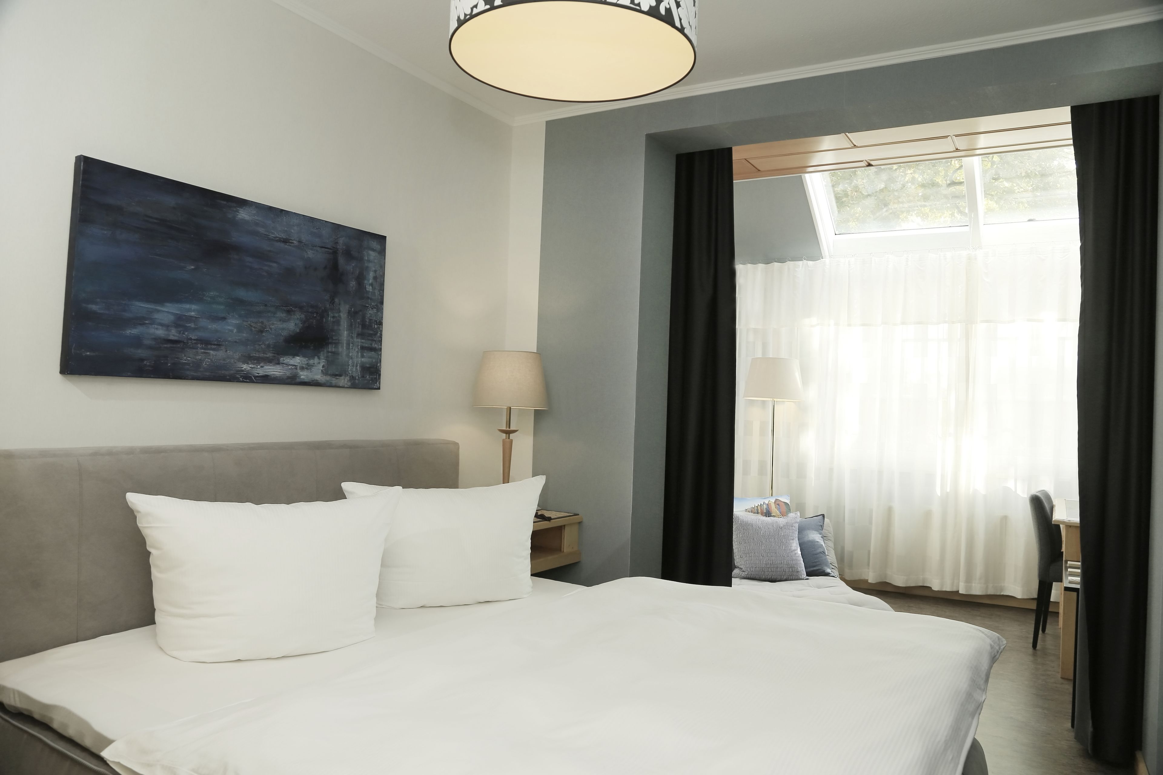 A hotel room with a large bed, a desk, a chair, a lamp, a painting, and a window with curtains.