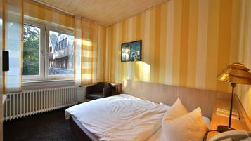 Une chambre d’hôtel avec un lit, une chaise, une table de chevet avec une lampe, un téléphone et une fenêtre donnant sur les bâtiments environnants.