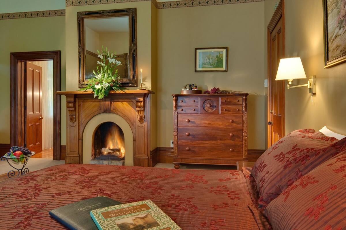 Een hotelkamer met een open haard, een houten dressoir, een bed met bloemenbedrukking en een ingelijst schilderij aan de muur.