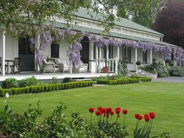 Una casa de estilo colonial blanco con un porche cubierto, mobiliario exterior y un césped bien cuidado con tulipanes rojos.