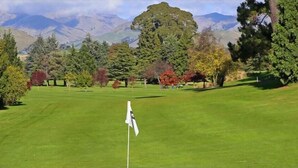 Ein Golfplatz mit Fahnenstange im Vordergrund, umgeben von Bäumen und Bergen im Hintergrund.