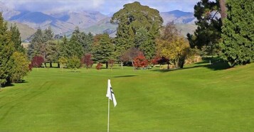 Un campo de golf con un marcador de bandera en primer plano, rodeado de árboles y montañas al fondo.