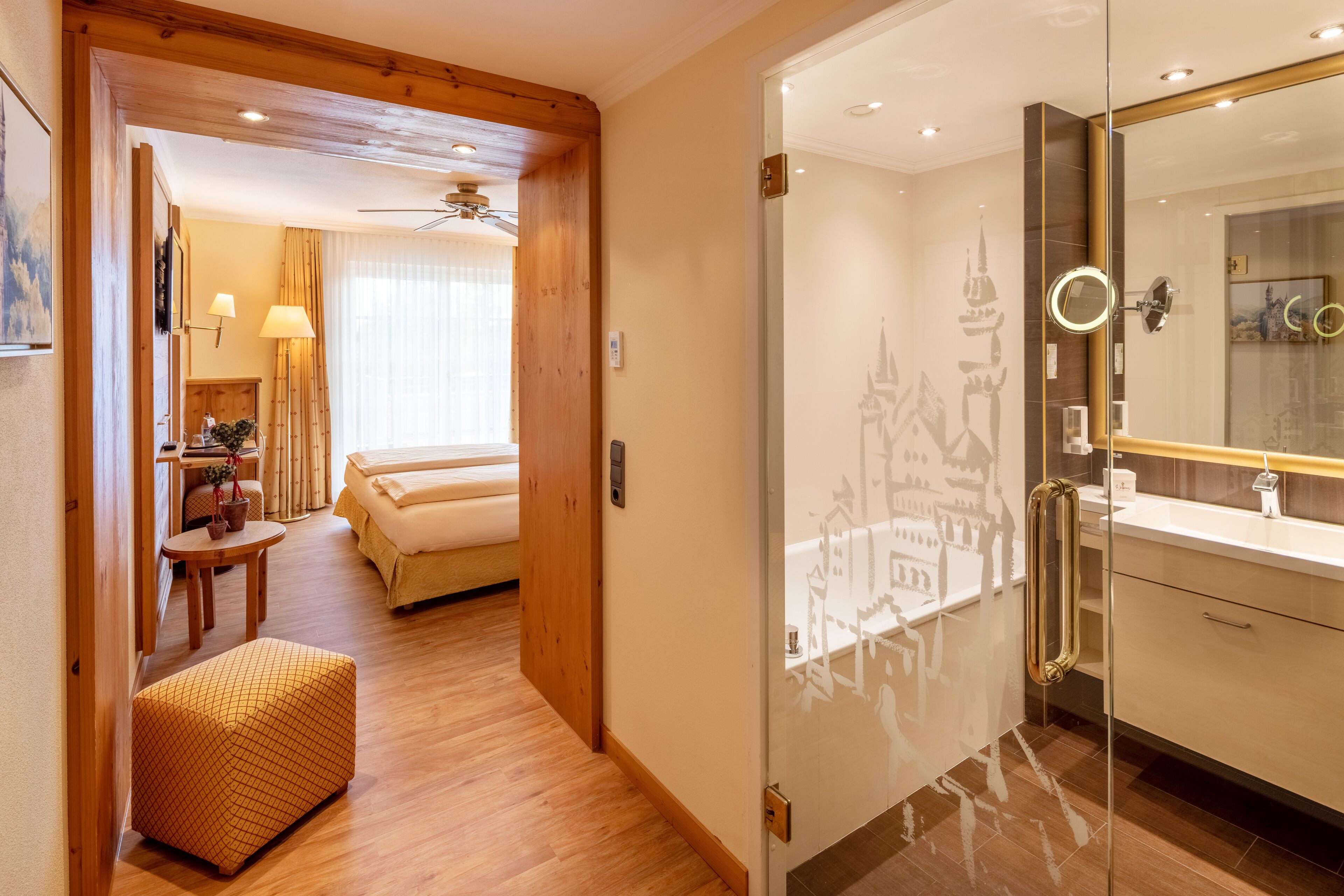 A hotel room with a wooden floor, a bed, a small table with a plant, a sofa, and a glass-enclosed bathroom with a mirror and sink.