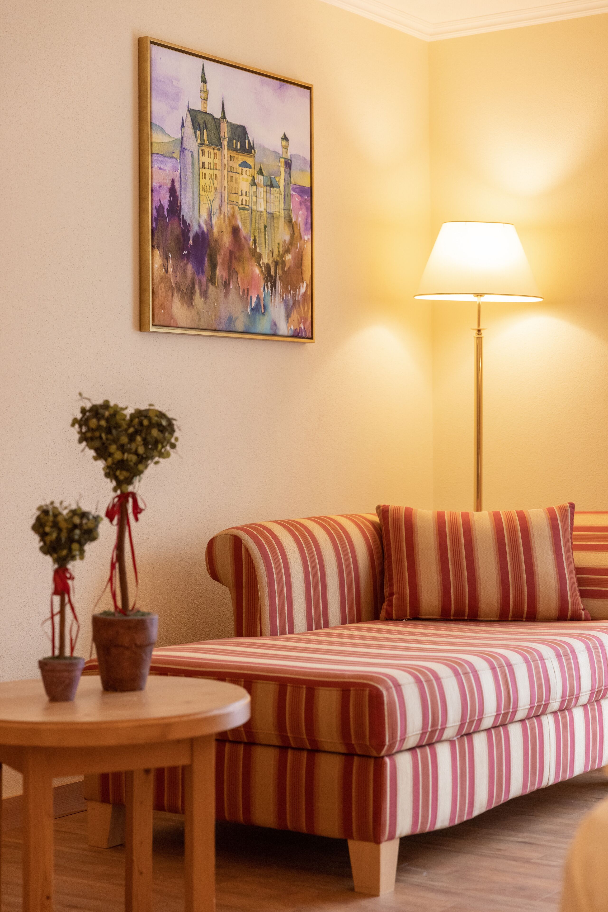 A room with a striped sofa, a round wooden table with potted plants, a floor lamp, and a painting of a castle on the wall.