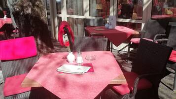 Un café al aire libre con sillas y mesas de mimbre cubiertas con manteles rojos, que cuenta con un cerramiento de cristal y un árbol al fondo.
