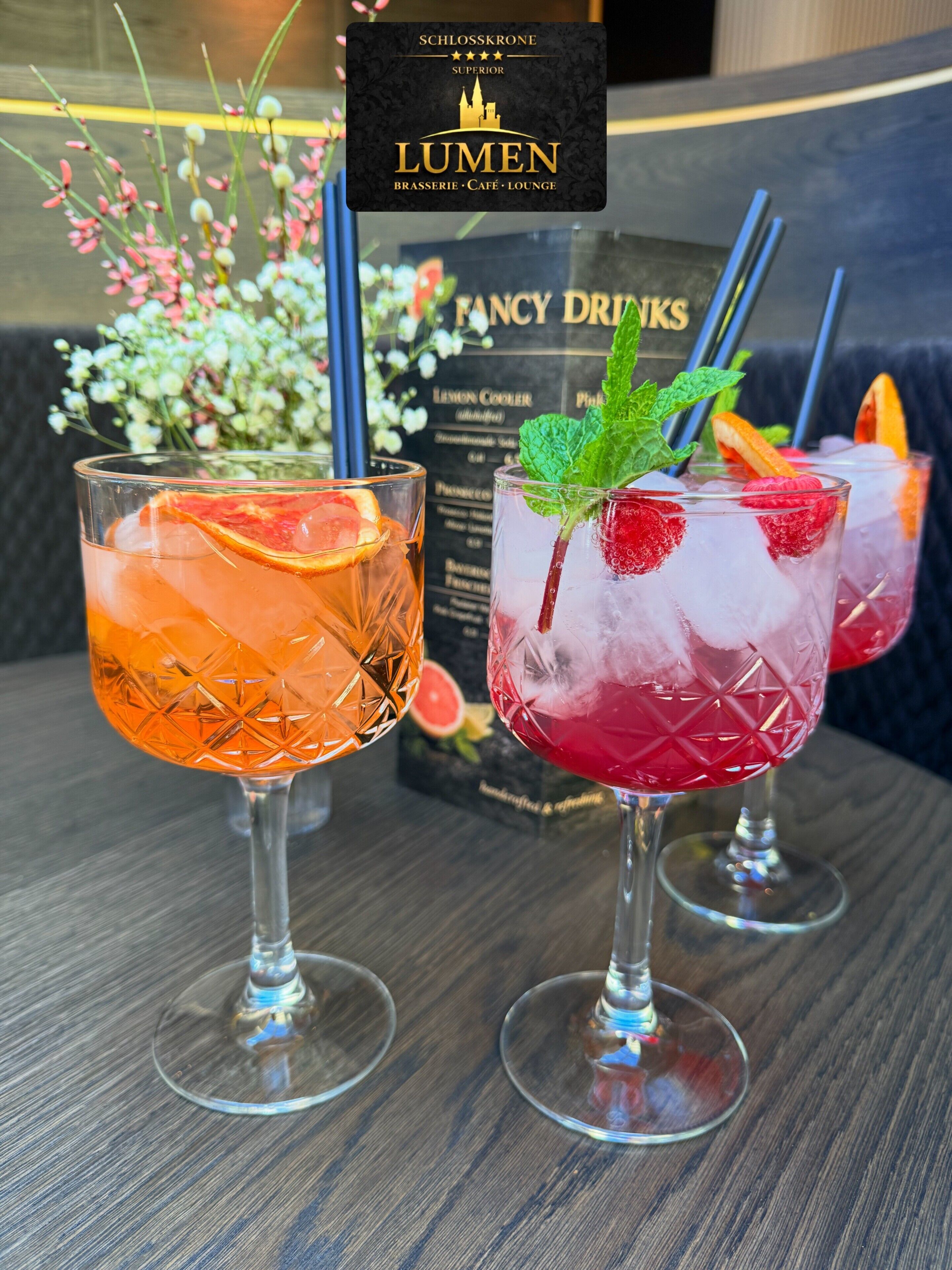 Dois cocktails com guarnições, um quadro de menu e uma placa com 'SCHLOSSKRONE LUMEN BRASSERIE CAFE LOUNGE'.