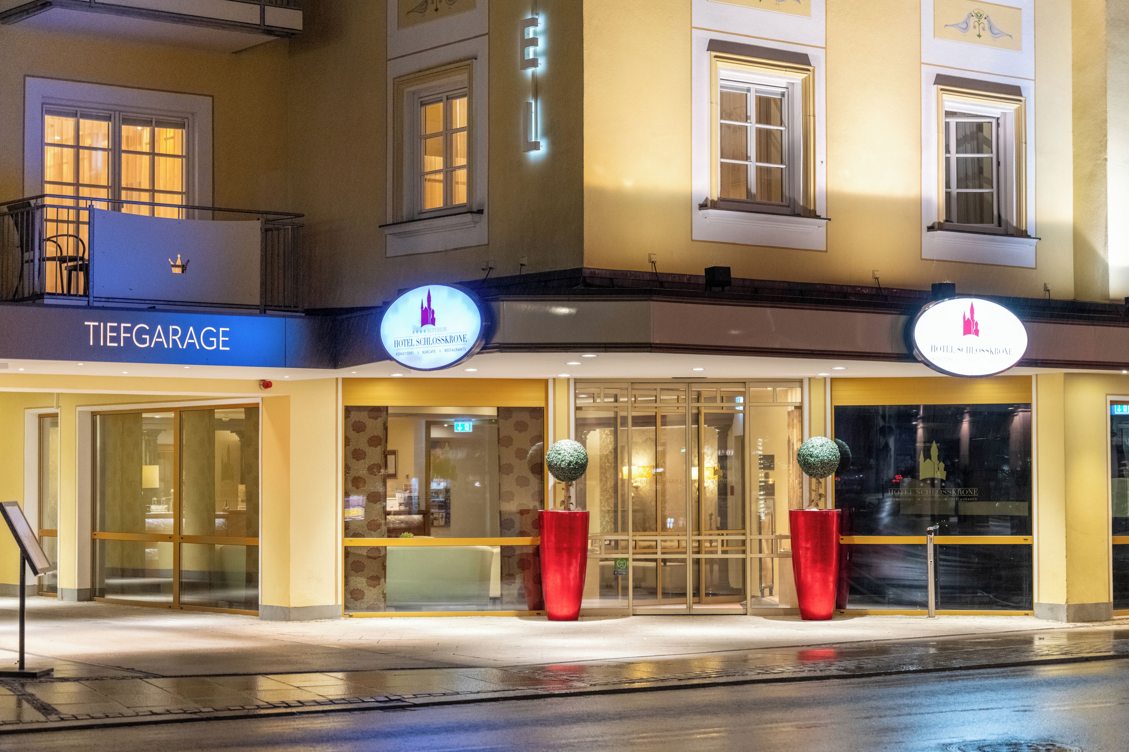 A hotel facade with a sign reading 'TIEFGARAGE HOTEL SCHLOSSBRUNE,' large windows, and two red planters.