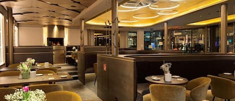 A modern restaurant interior with wooden ceiling panels, round tables, and yellow ambient lighting.