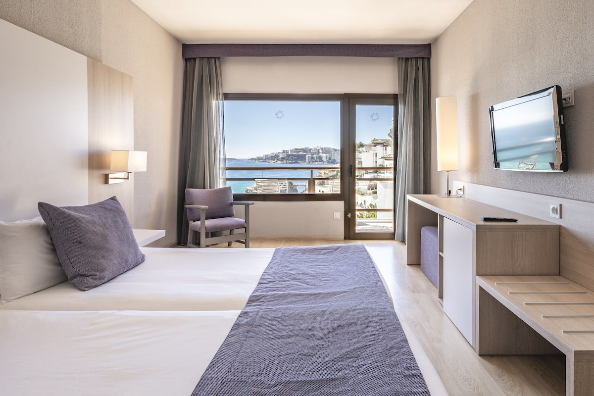 Habitación de hotel con cama, escritorio, silla, televisión y vista al mar.