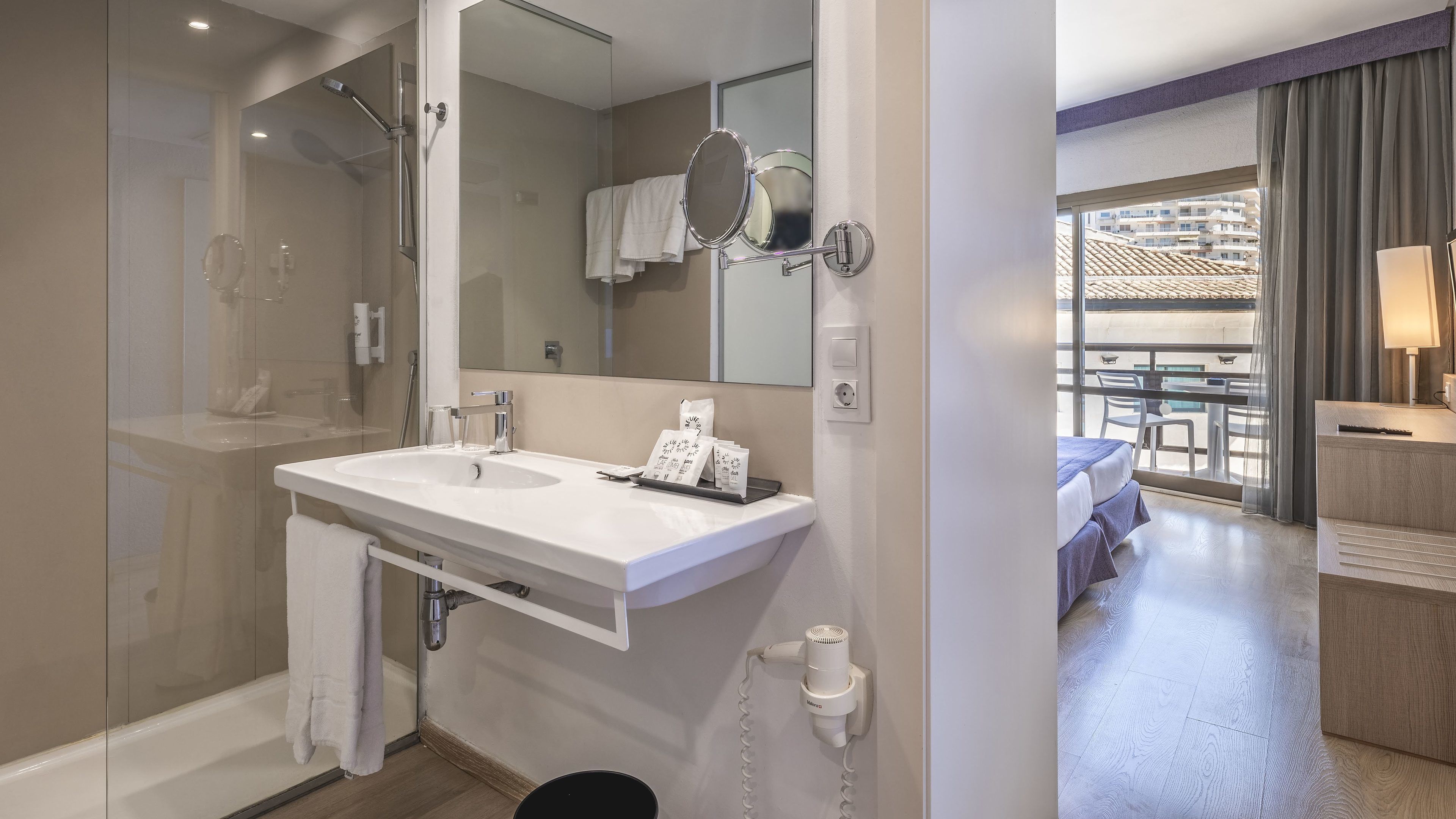 A modern bathroom with a large mirror, a shower, and a vanity with a sink. There is a small table with toiletries and a towel hanging on the wall.