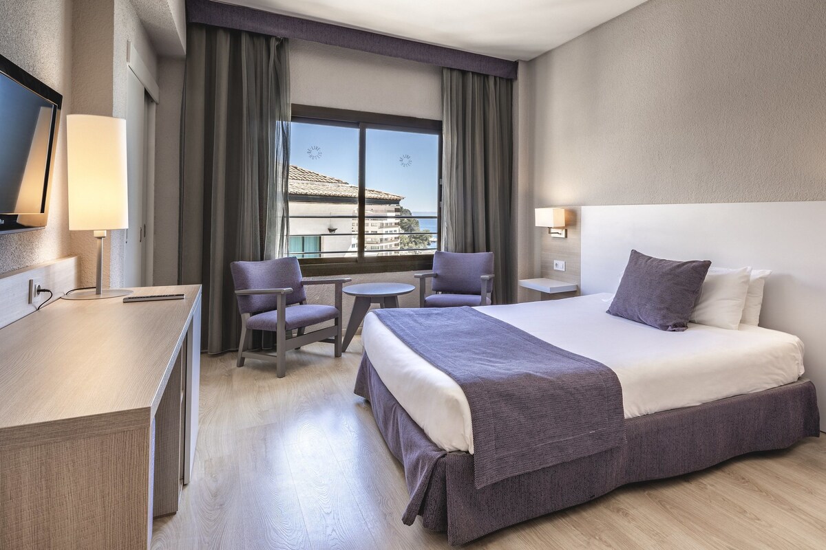 Ein Hotelzimmer mit Bett, Schreibtisch, Stuhl, kleinem Tisch und einem Fenster mit Vorhängen.