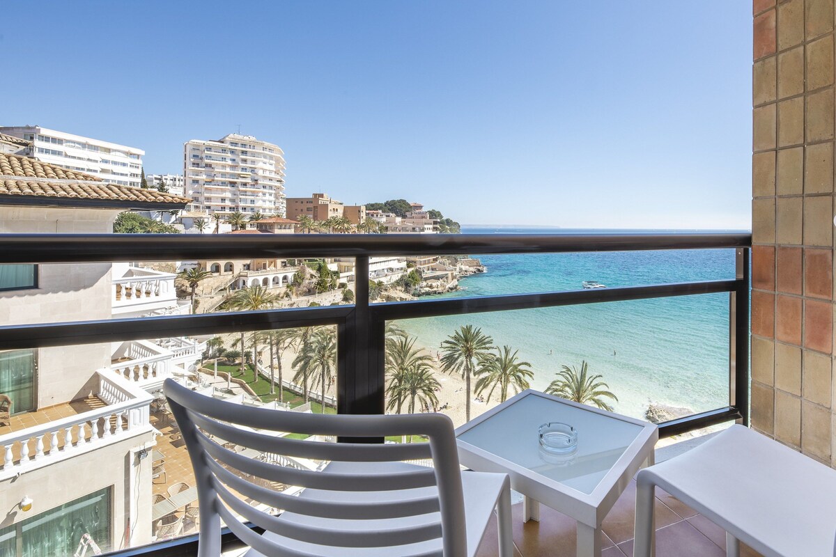 Ein Balkon mit weißen Stühlen und einem Tisch, der Blick auf einen Strand und eine Stadt bietet.