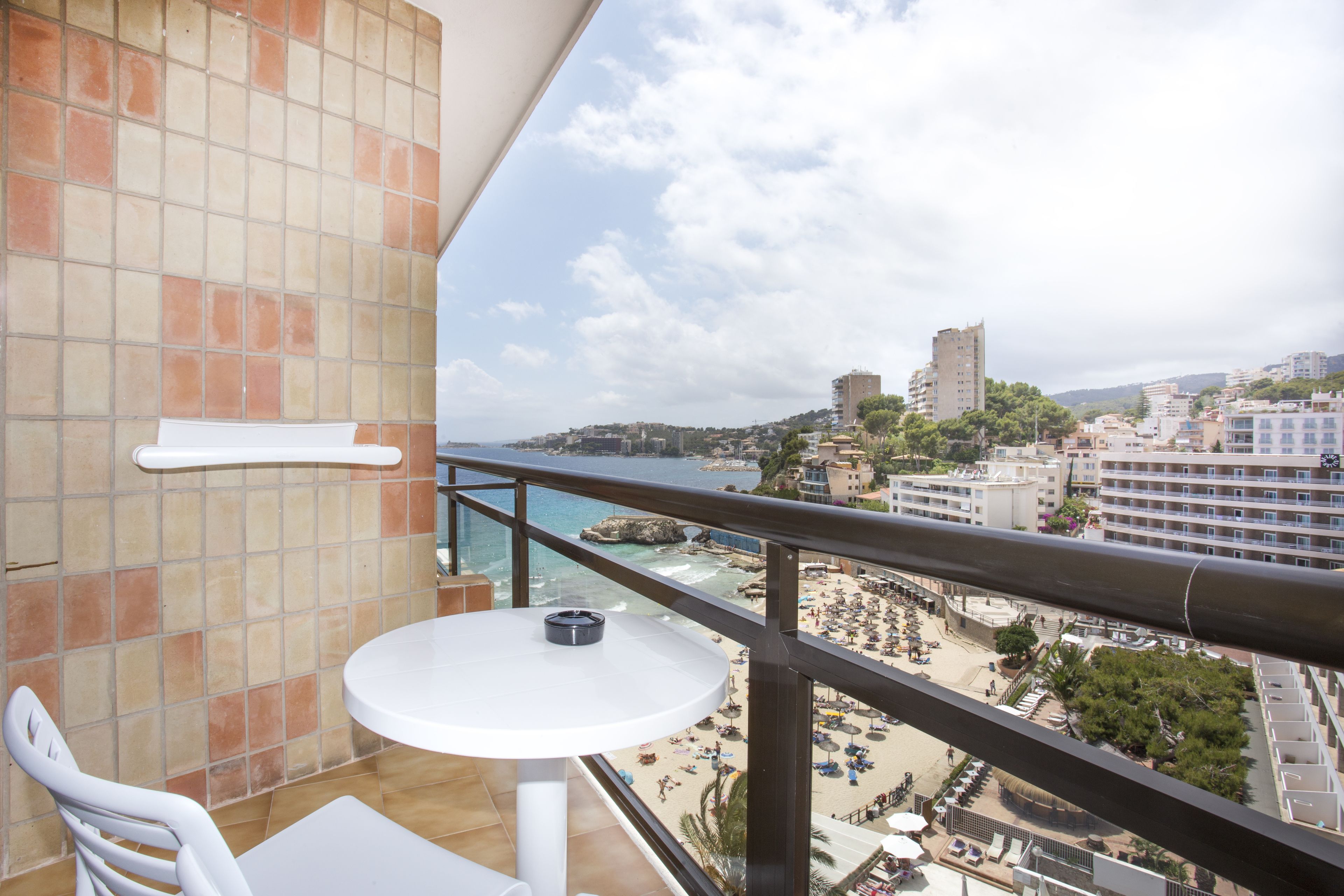 Un balcón con una mesa y silla blancos, con vista a una playa con sombrillas y gente, y edificios al fondo.