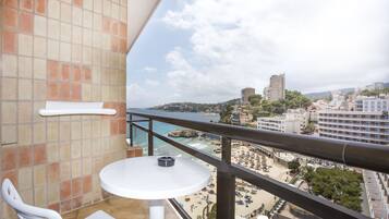 Un balcón con una mesa y silla blancos, con vista a una playa con sombrillas y gente, y edificios al fondo.
