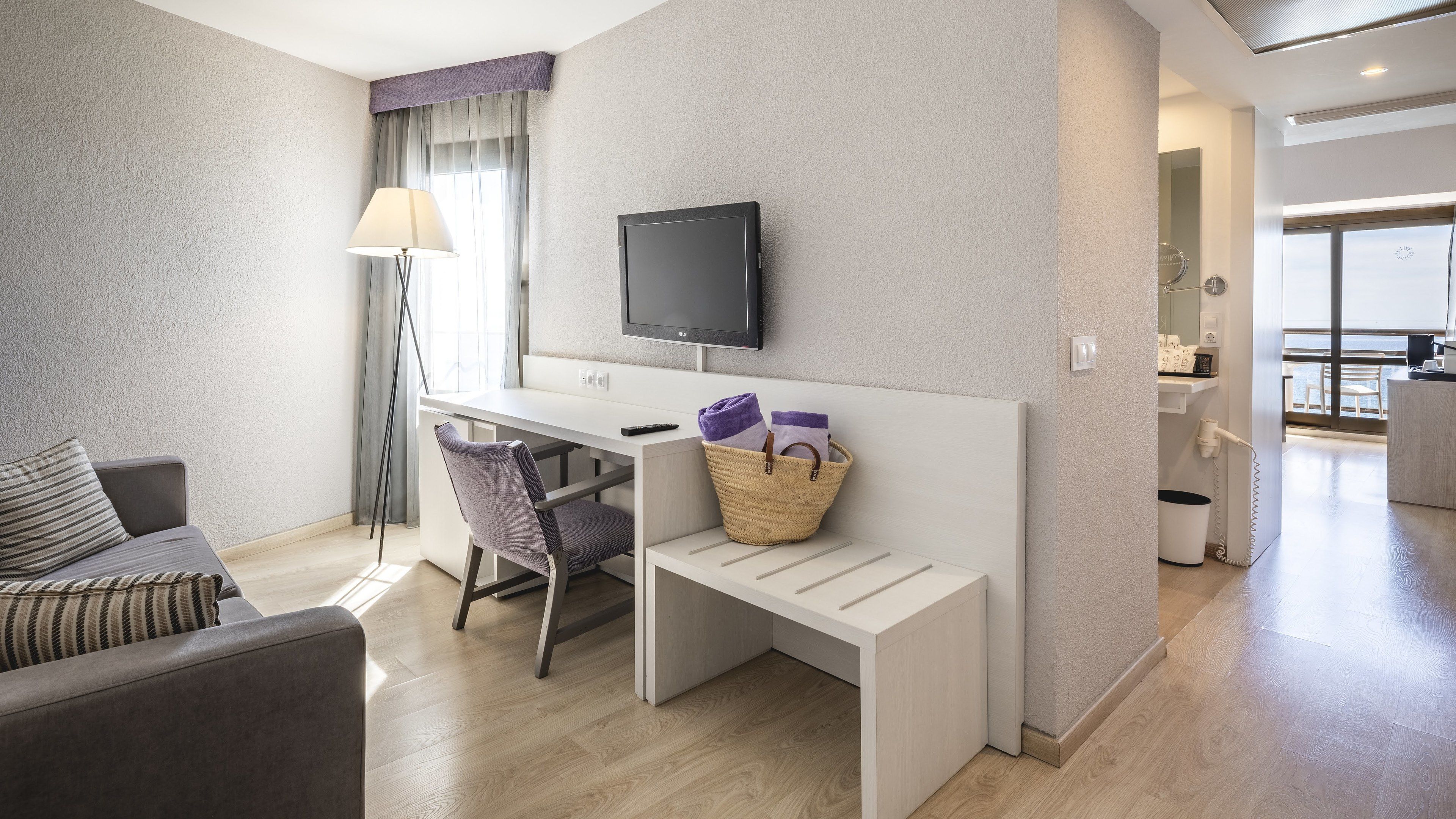 A modern hotel room with a flat-screen TV, a desk, a chair, a sofa, and a small table.