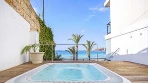 Un jacuzzi rectangular en una terraza de madera, con vista al mar y un edificio blanco.