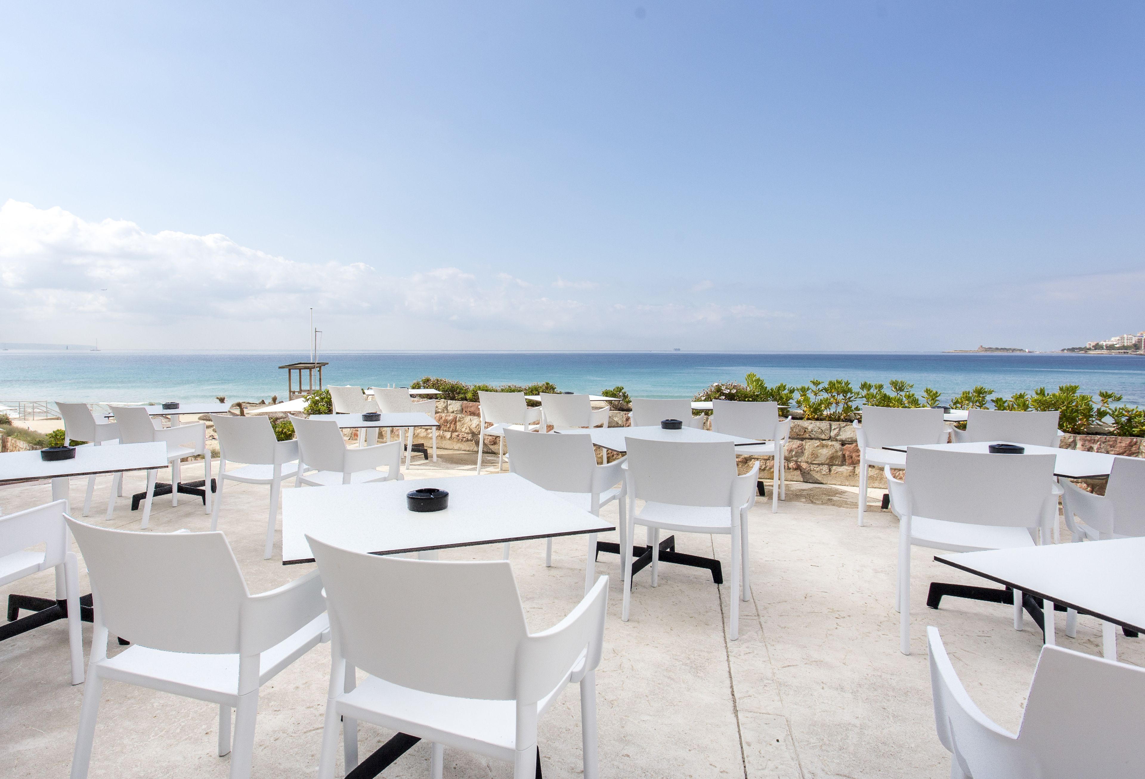 An outdoor seating area with white tables and chairs, overlooking a beach and the sea.