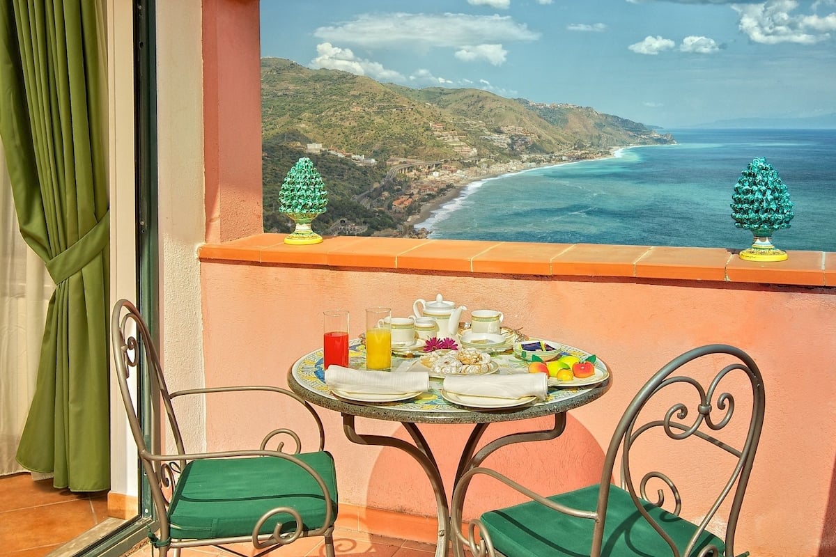 A balcony with a table set for breakfast, overlooking a coastal landscape with trees and a view of the sea.