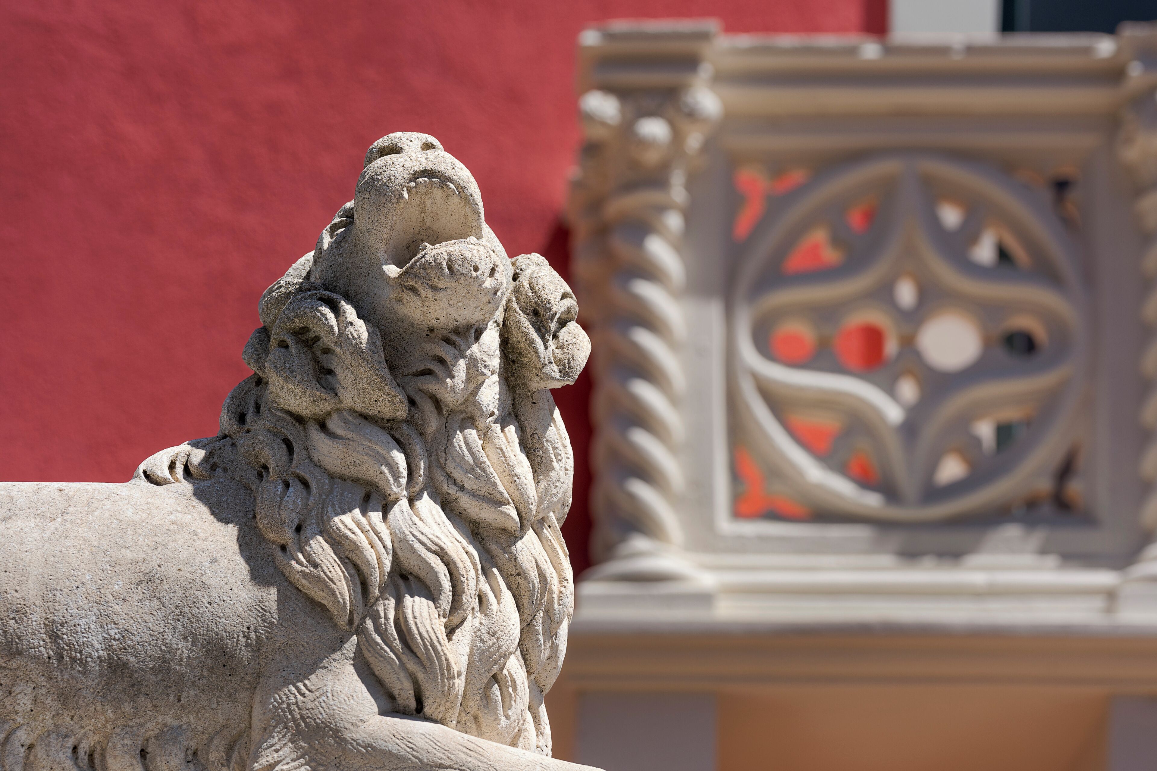 A stone lion statue with intricate carvings, set against a red wall and a decorative metal fence.