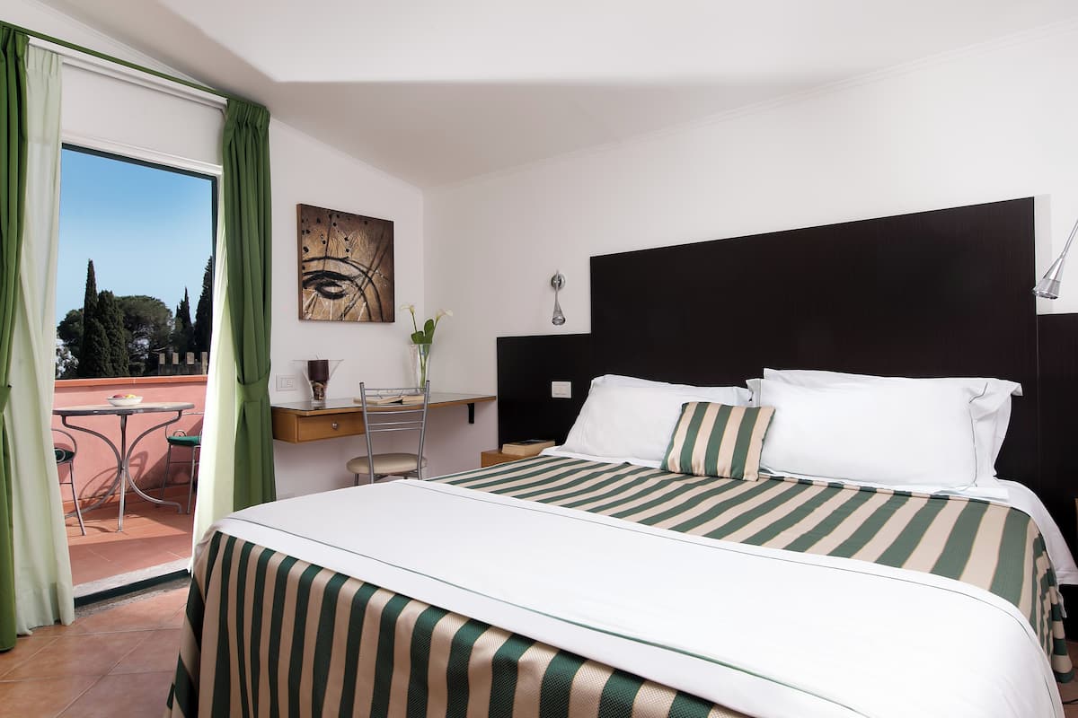 A hotel room with a large bed, a desk with a chair, a view of the outdoors, and a painting on the wall.