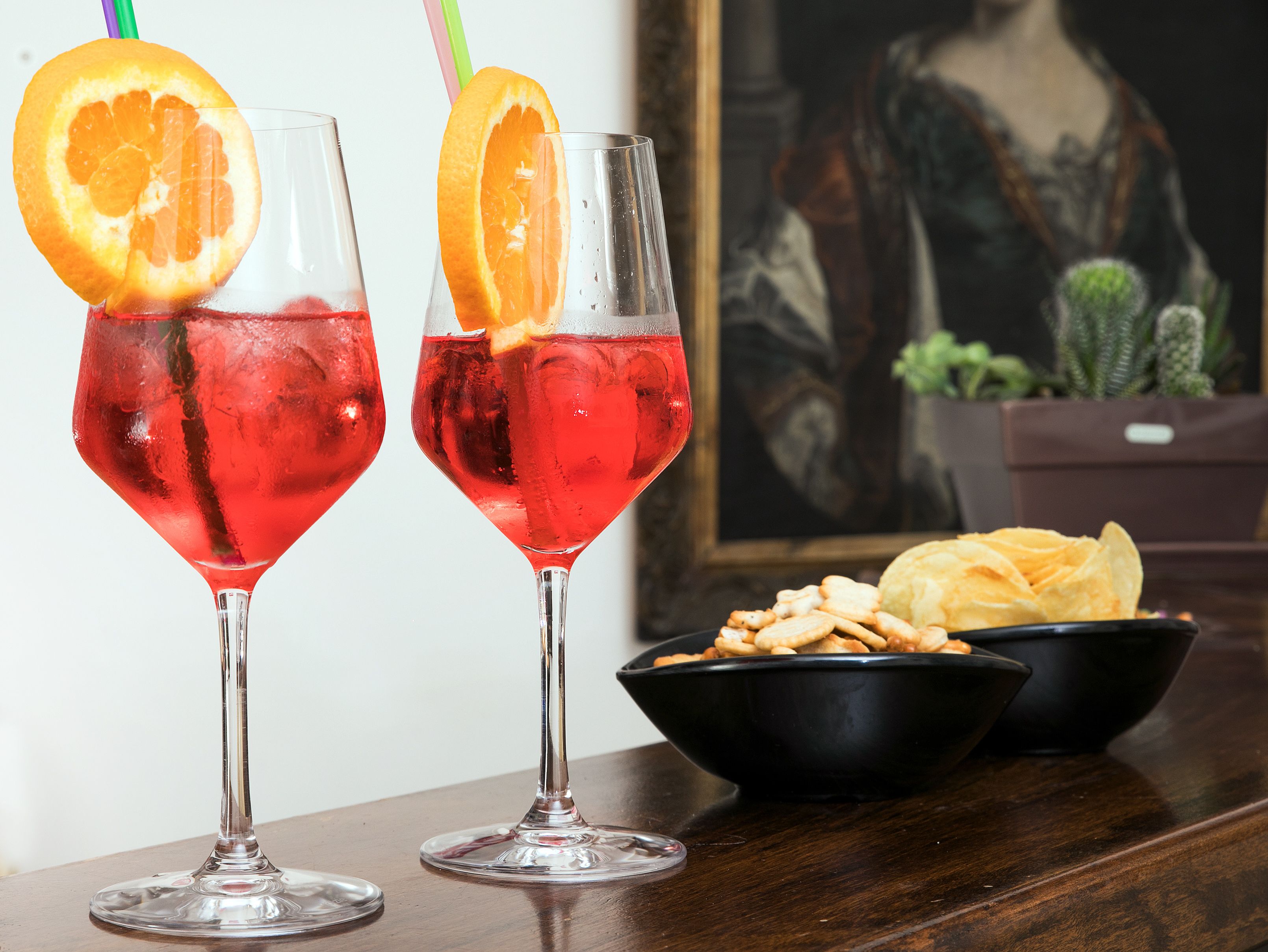 Two glasses of red beverage with orange slices and straws, a bowl of snacks, and a painting in the background.