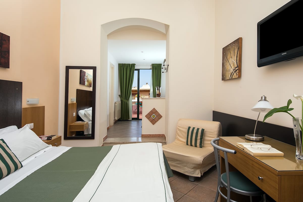 A hotel room with a bed, a desk, a chair, a TV, and a view of the outside.