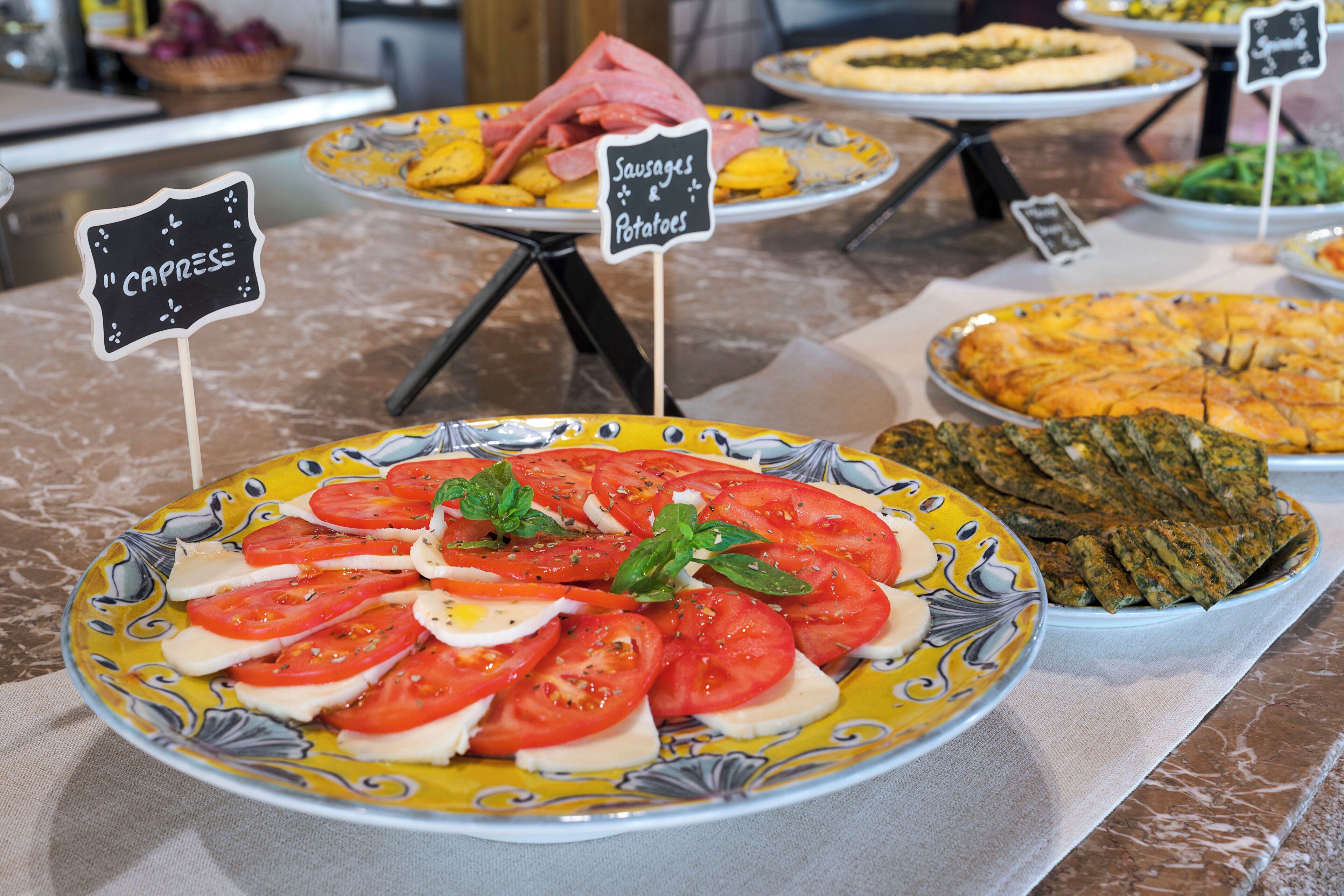 Ein reichhaltiges Buffet mit verschiedenen Speisen, darunter ein Teller mit Tomaten und Mozzarella, sowie Schilder, die Essensoptionen wie 'Sausages & Potatoes' und 'Caprese' bezeichnen.