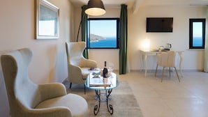 A hotel room with a large window, a flat-screen TV, two armchairs, a small table with a bottle and glass, and a desk with a laptop and a cup.