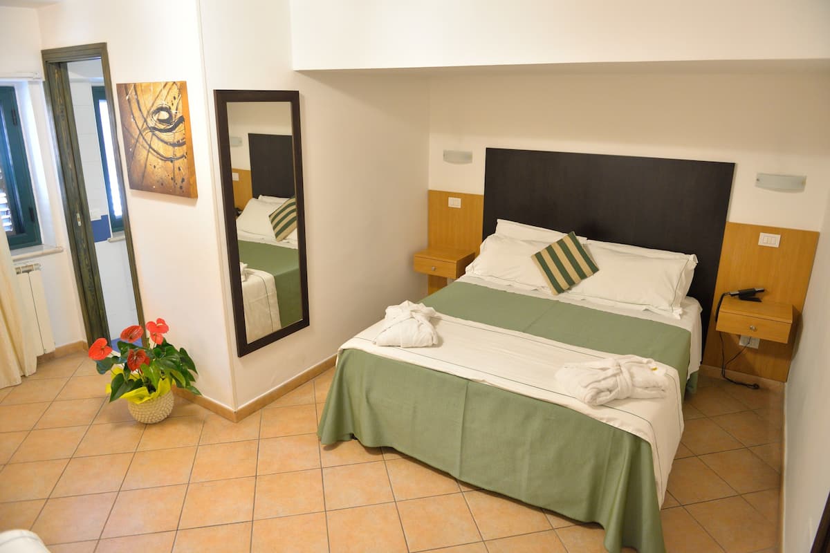 A hotel room with a bed, a mirror, a nightstand, a bedside table, and a potted plant.