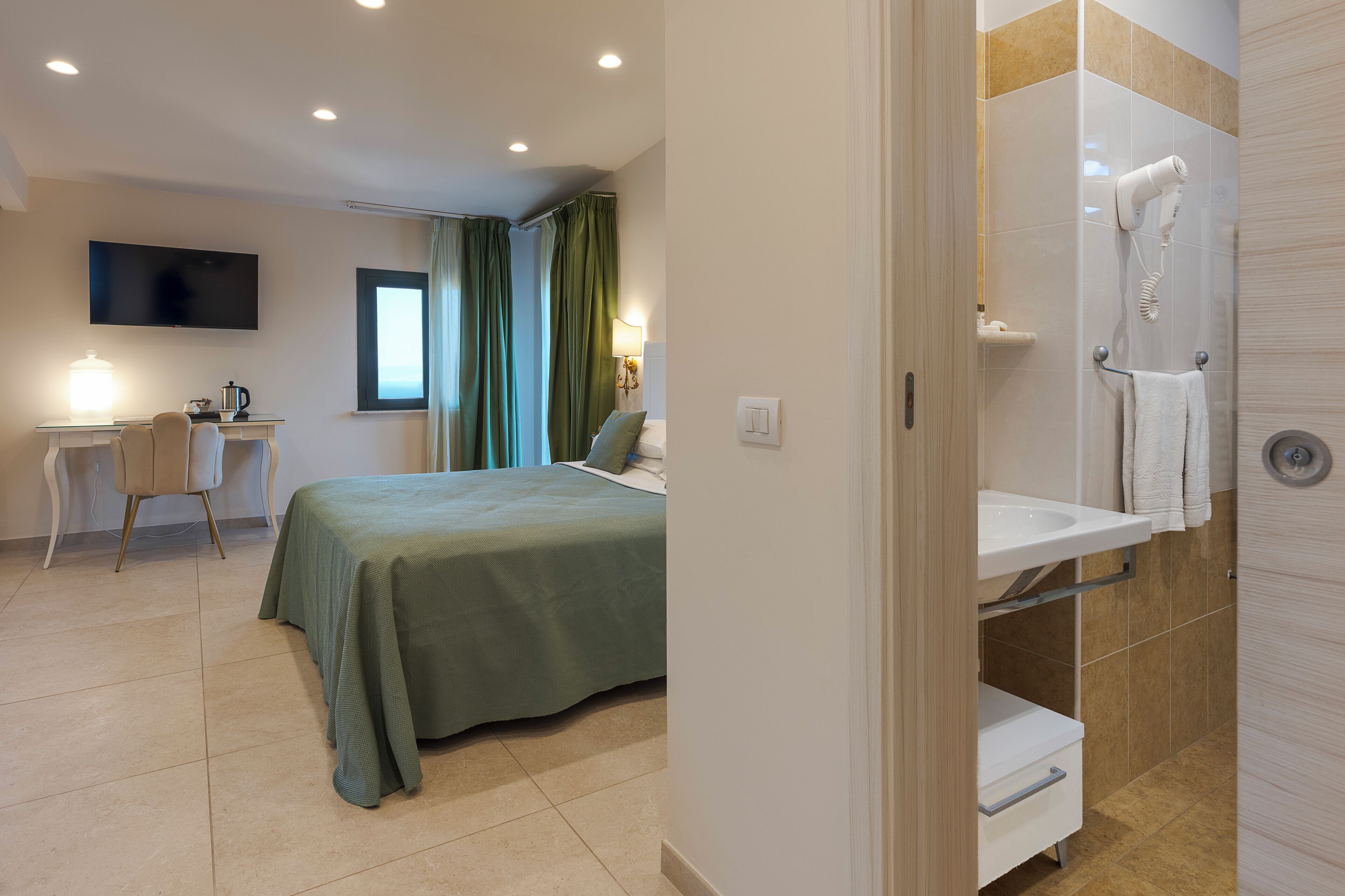 A hotel room with a bed, a desk with a chair, a TV, a shower, and a bathroom sink.