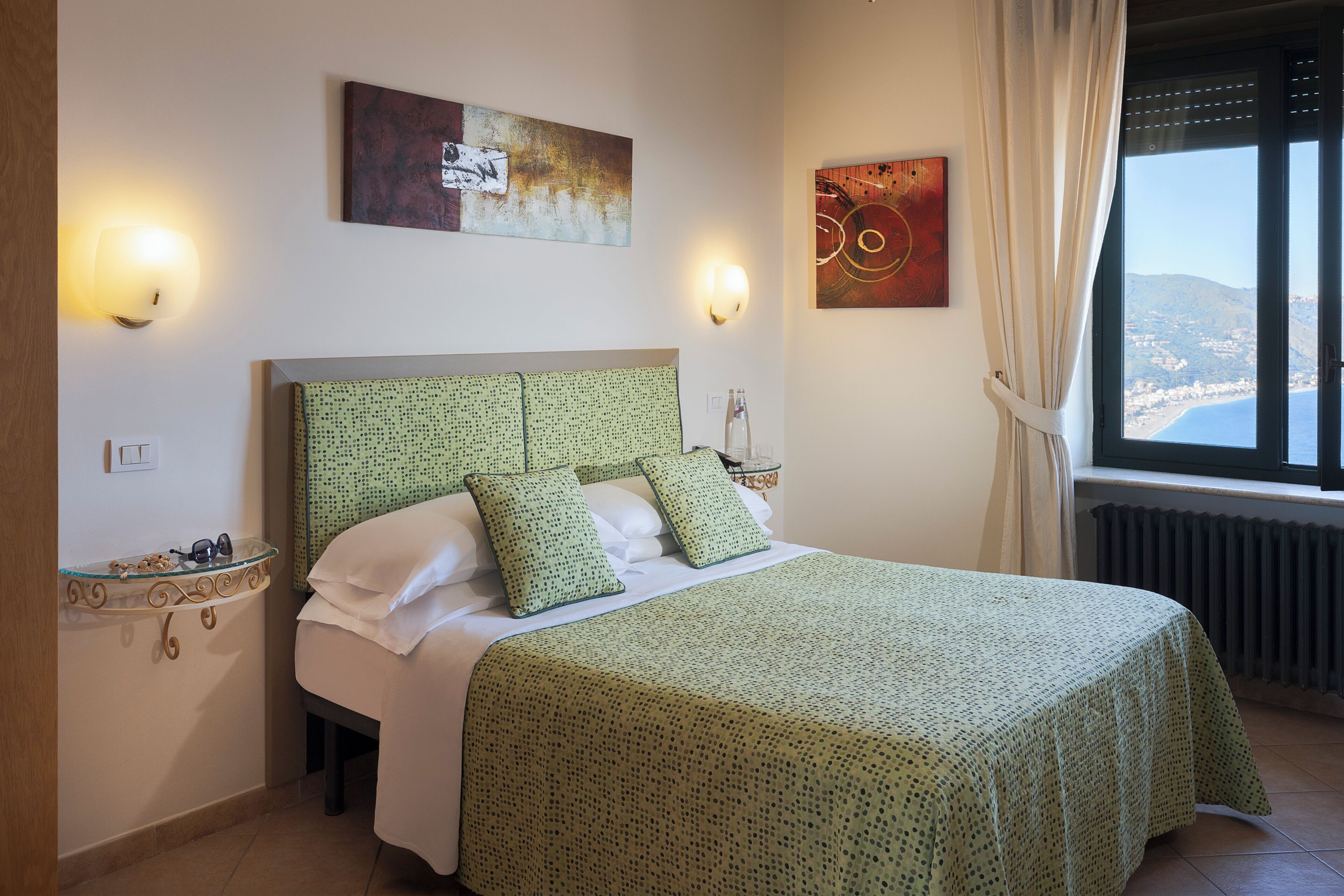 A bedroom with a bed, two wall-mounted lamps, a window with curtains, and artwork on the walls.