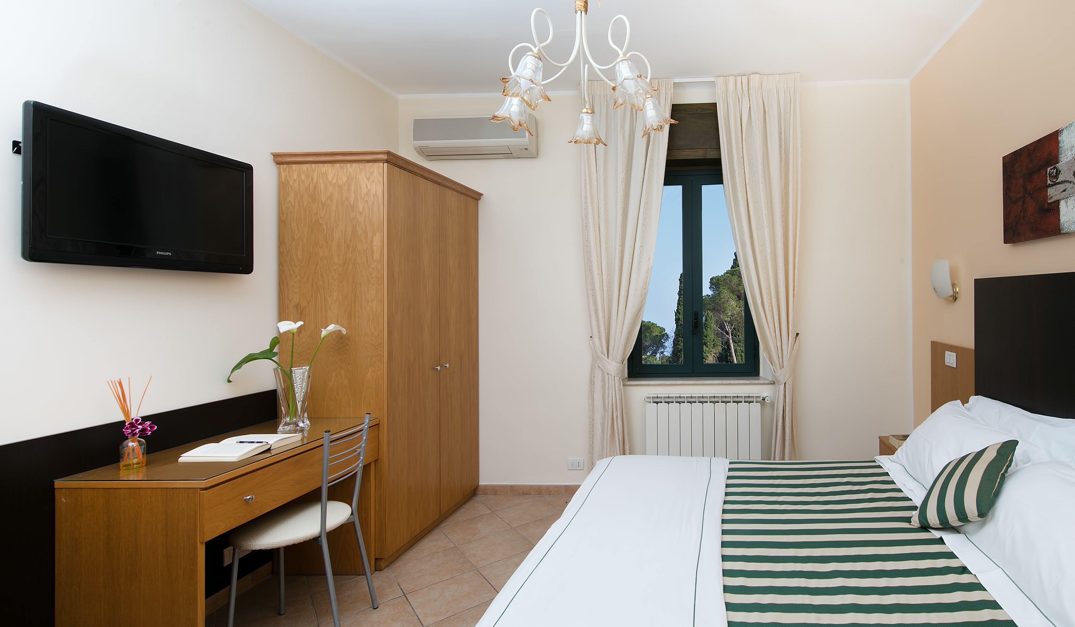 Une chambre d’hôtel comprenant un lit, un bureau, une chaise, une télévision, une armoire, une fenêtre avec des rideaux et un radiateur.