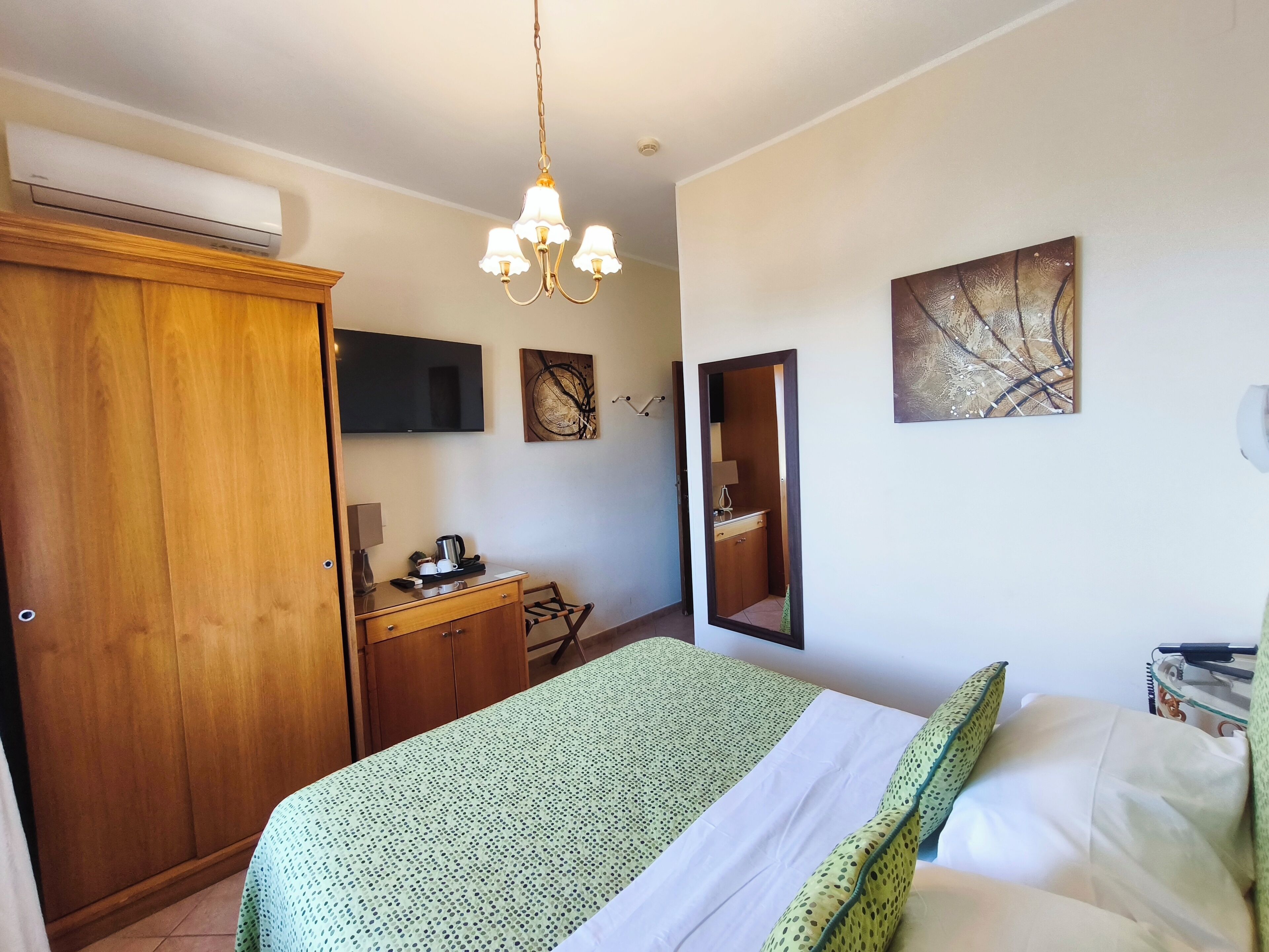 A bedroom with a bed, a wooden wardrobe, a desk with a chair, a television, and artwork on the walls.