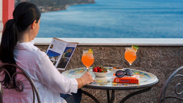 A person reading a book at a table with a view of the sea, two drinks, a bowl of fruit, and sunglasses.