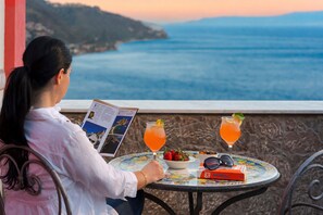 A person reading a book at a table with a view of the sea, two drinks, a bowl of fruit, and sunglasses. - Hotel La Pensione Svizzera (Taormina)
