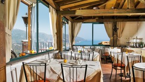 A restaurant with multiple tables and chairs, large windows with a view of the sea, and a rustic wooden ceiling.