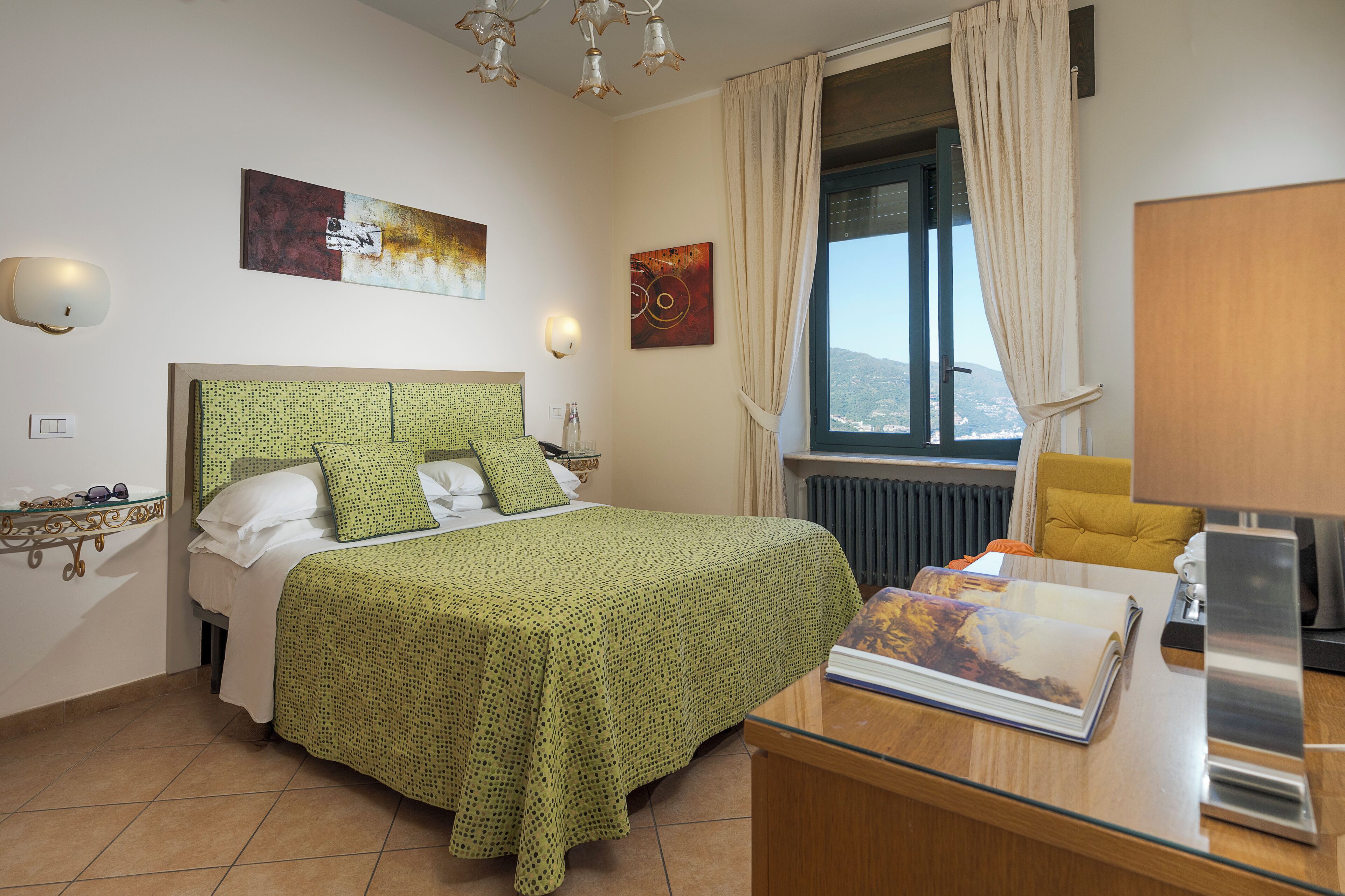 Un dormitorio con cama, una mesita de noche con un libro, una ventana con cortinas y obras de arte en las paredes.