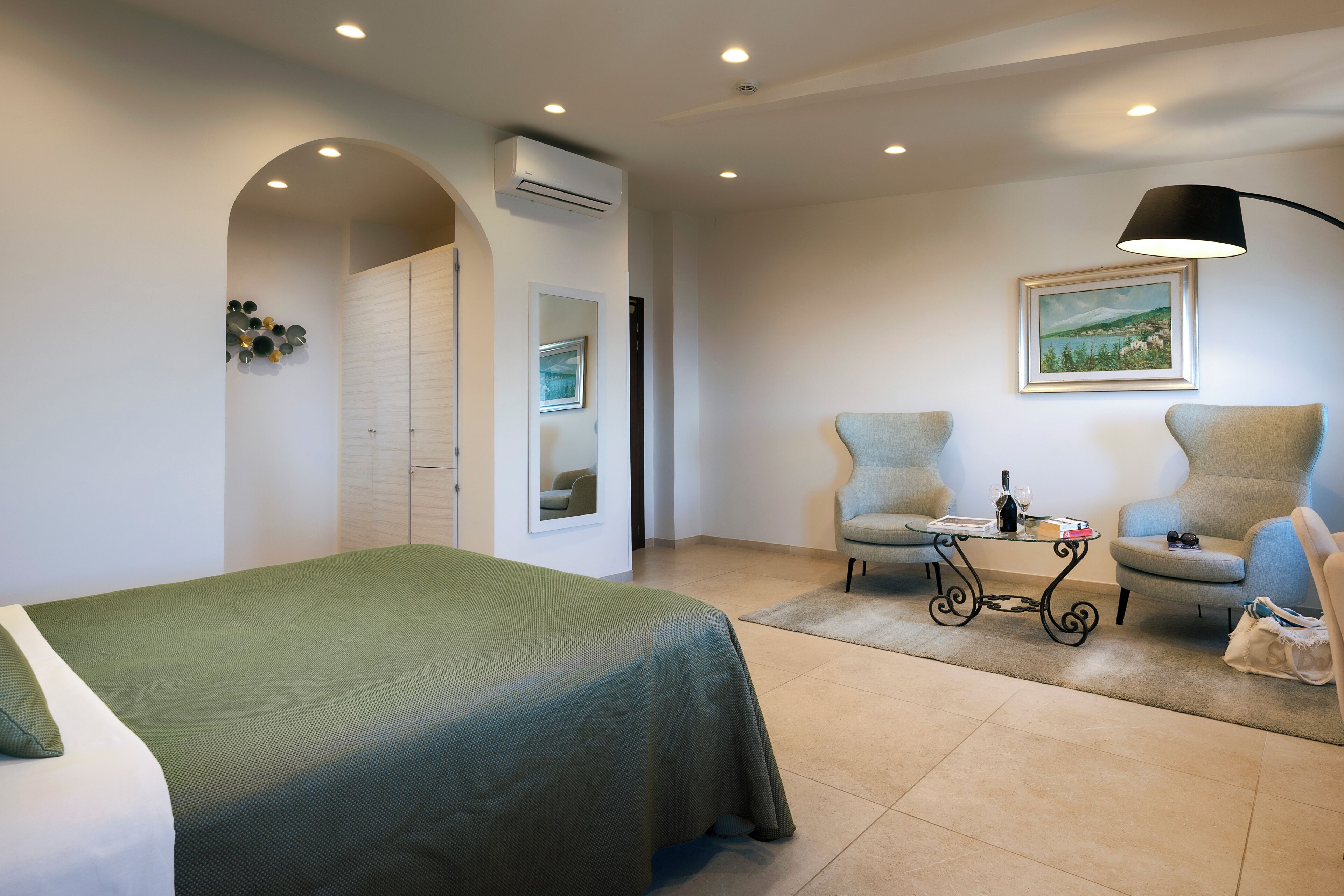 Quarto de hotel com uma cama, duas poltronas, uma mesa com abajur, um quadro na parede e um ar condicionado.