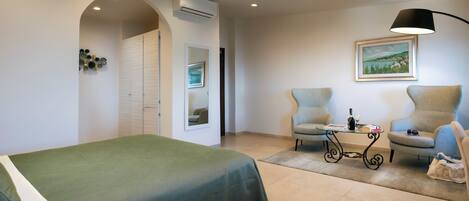 Quarto de hotel com uma cama, duas poltronas, uma mesa com abajur, um quadro na parede e um ar condicionado.