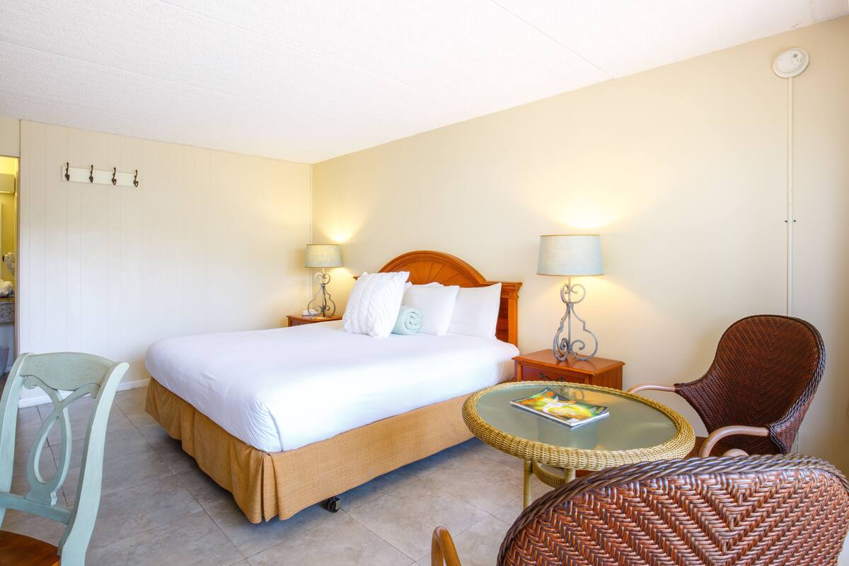 Una habitación de hotel con una cama grande, dos mesitas de noche con lámparas, un sillón de mimbre, una pequeña mesa redonda con una revista y una silla con asiento de fibra.
