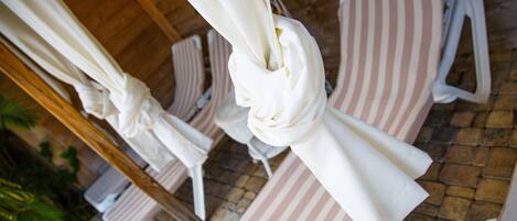 Un conjunto de sillones reclinables a rayas con toldos blancos, dispuestos en fila sobre un patio con baldosas.