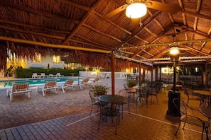 An indoor pool area with lounge chairs and tables, a thatched roof, and string lights.
