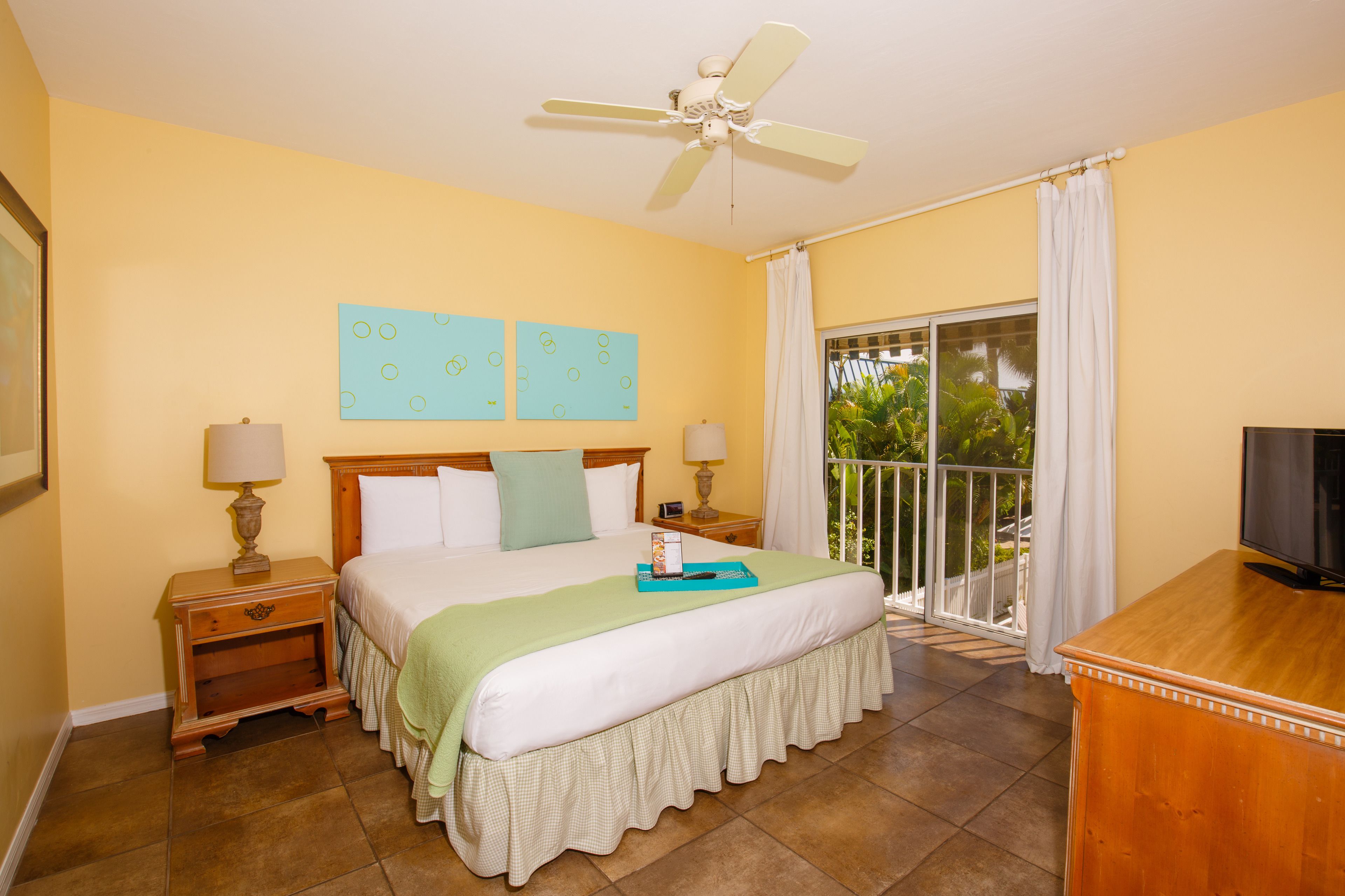 A hotel room with a bed, two bedside tables with lamps, a ceiling fan, a TV, and a balcony with curtains.
