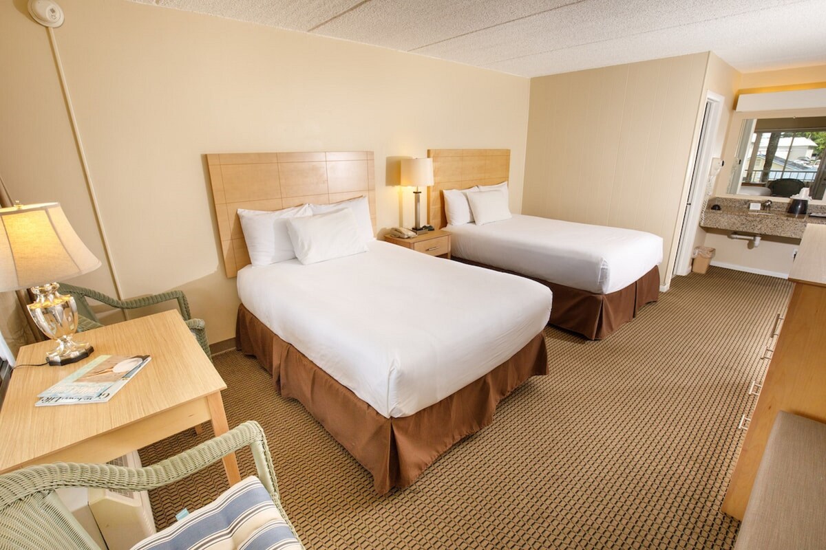 Quarto de hotel com duas camas, uma escrivaninha com luminária, uma cadeira e uma mesinha.
