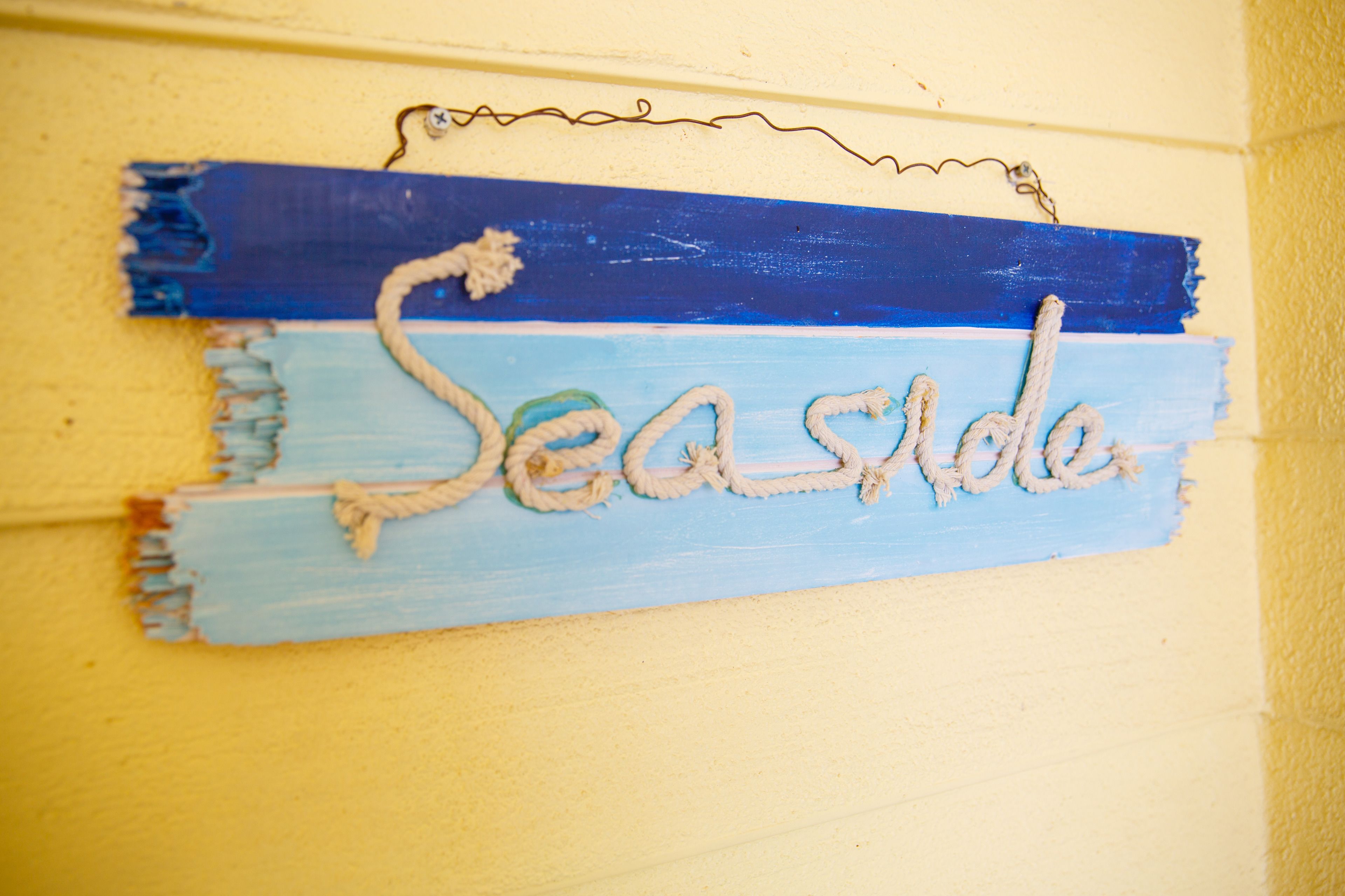 A sign with the word "Seaside" painted in a blue and beige color scheme, mounted on a yellow wall.